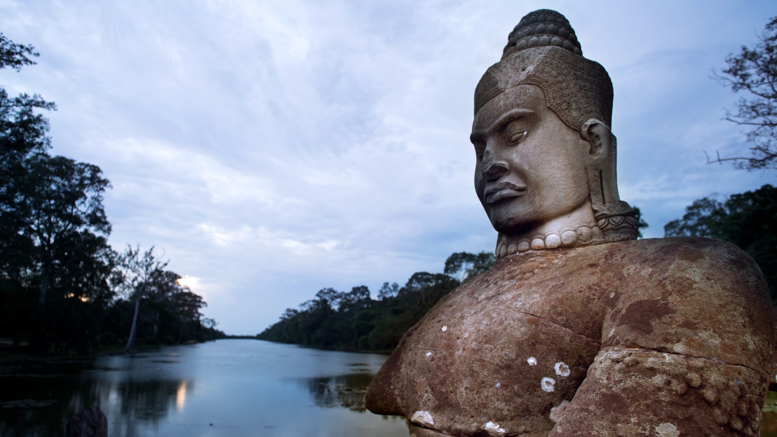 Stone sentinel statue at dusk along a still temple moat